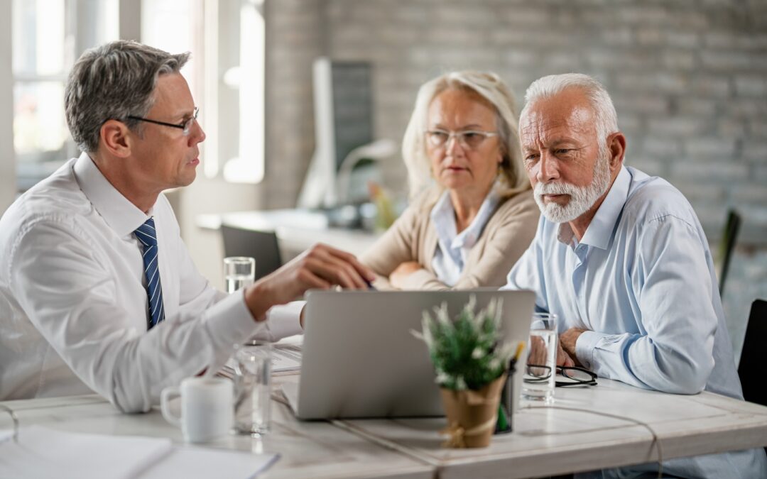 Un conseiller financier discute avec un couple de seniors autour d’un ordinateur portable dans un bureau lumineux, probablement en train de parler d’épargne ou de retraite.