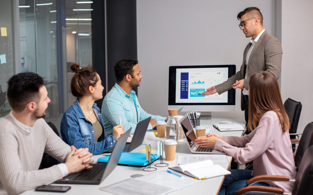 Un homme en costume présente des graphiques financiers sur un écran d'ordinateur à une équipe de jeunes professionnels réunis autour d'une table de bureau